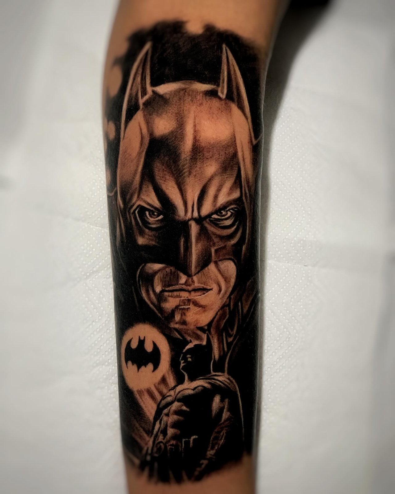Tatuagem realista no antebraço: rosto do Batman com símbolo do morcego e silhueta na cidade