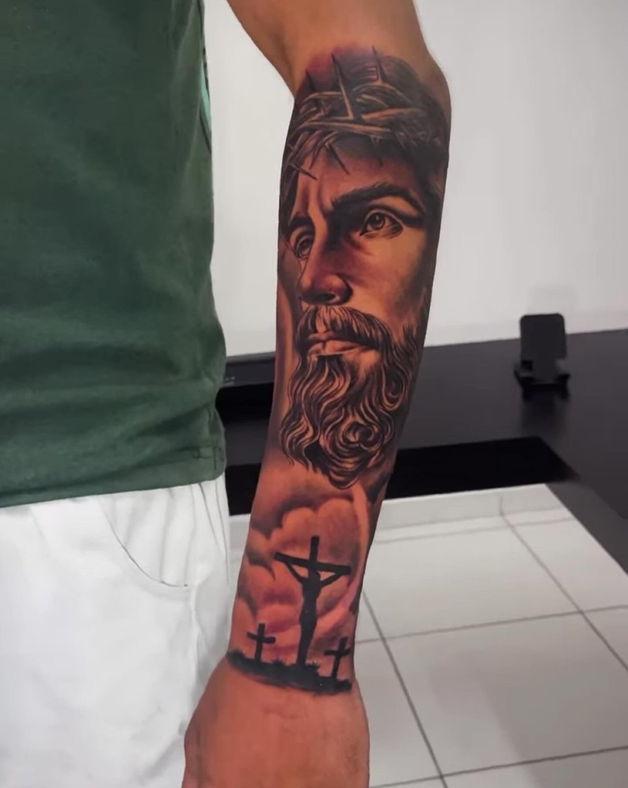 Tatuagem realista no antebraço: rosto de Jesus com coroa de espinhos sobre as três cruzes do Calvário