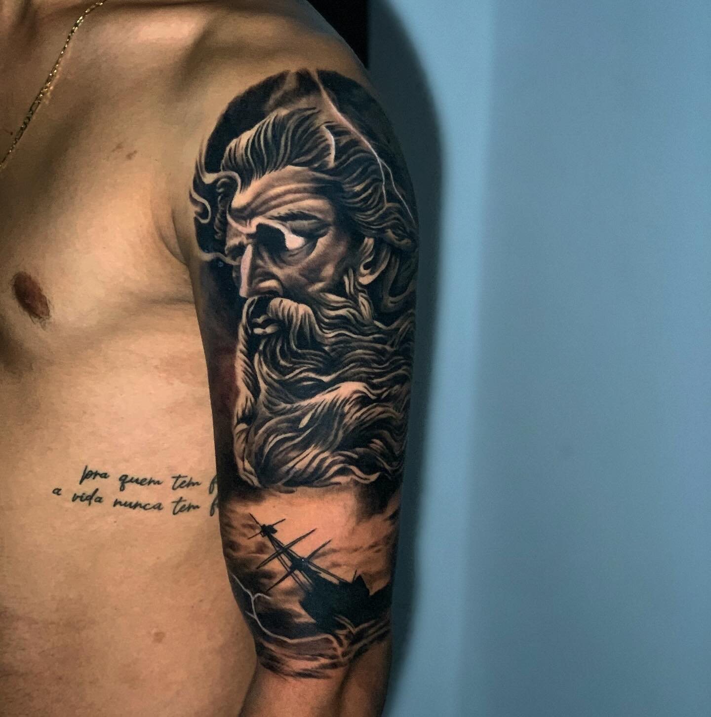 Tatuagem realista no ombro e braço: rosto de Zeus com barba e navio em mar revolto